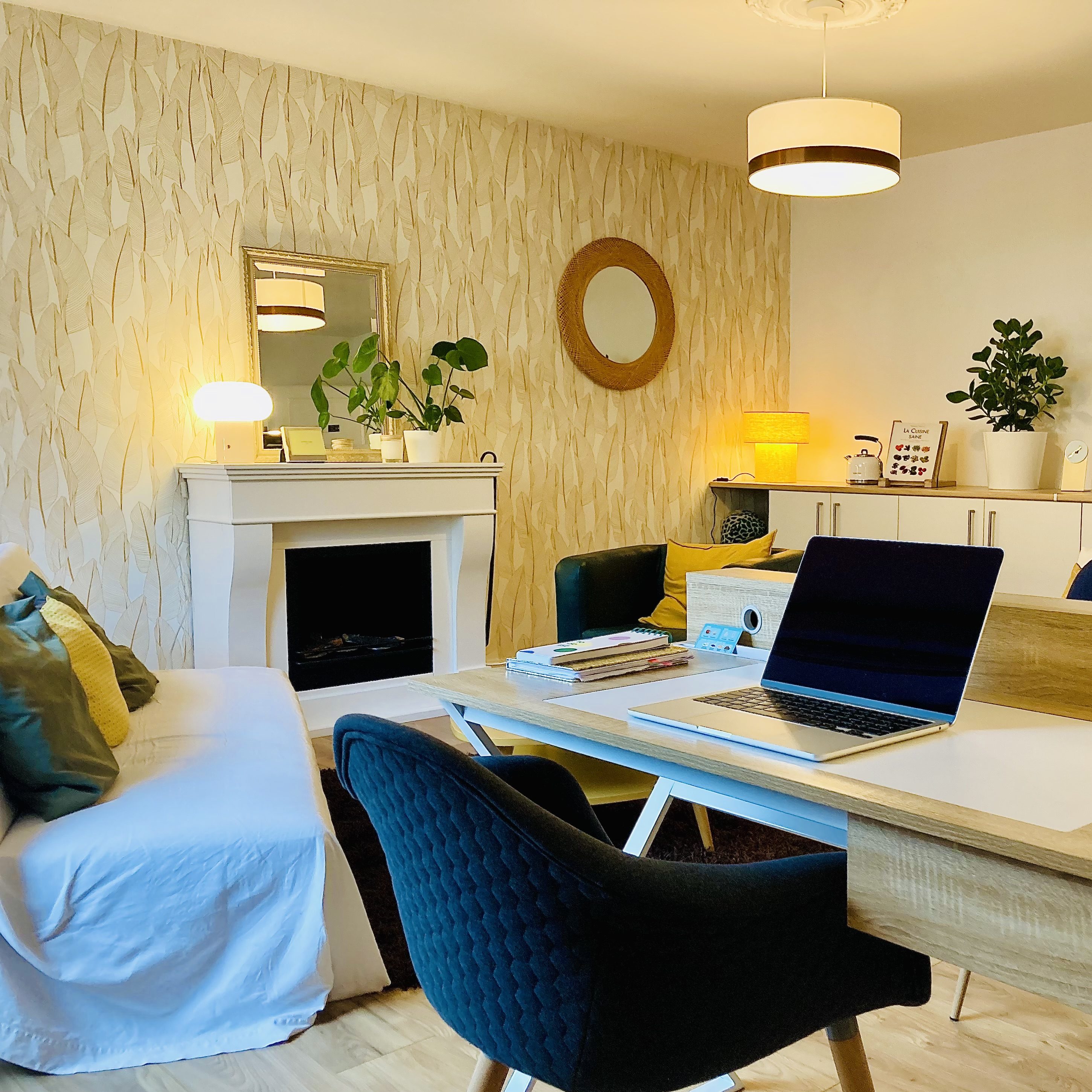 Un salon moderne avec un canapé blanc, une cheminée, des plantes sur une étagère, et un bureau en bois avec un ordinateur portable.